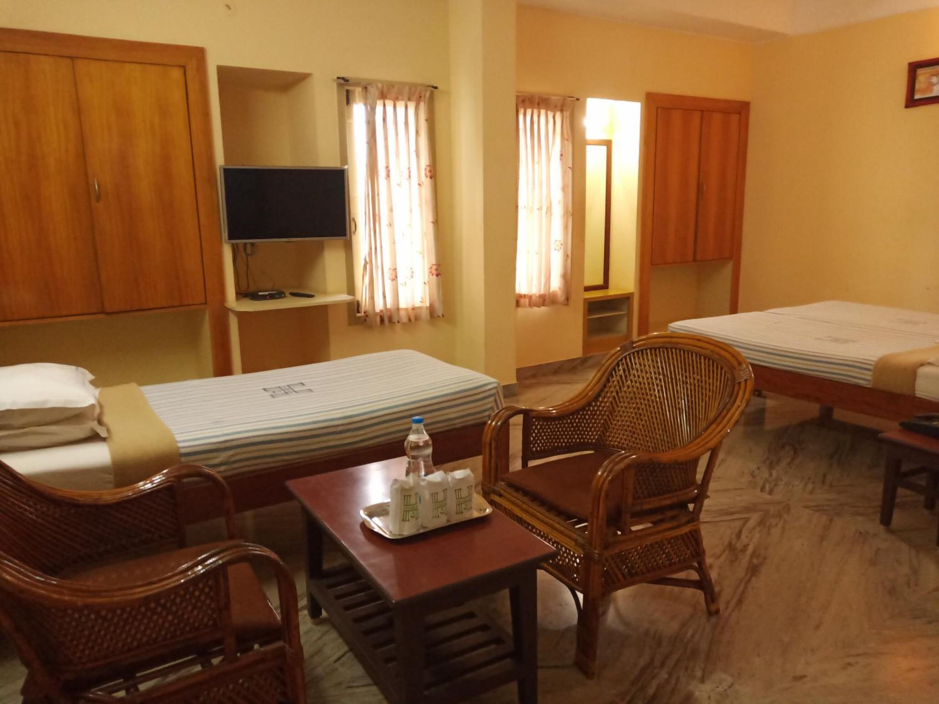 A hotel room with two single beds, a wooden desk with a chair, a television, and a small table with a bottle and glasses.