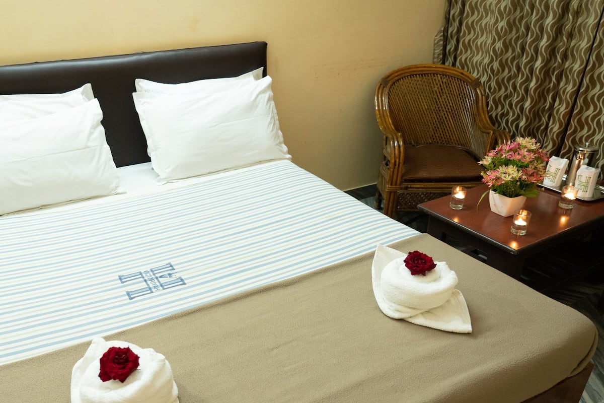 Una habitación de hotel con cama, almohadas, una mesita de noche con un florero y una silla.