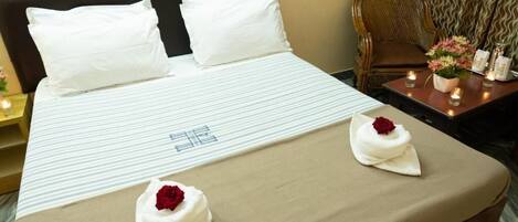 Habitación de hotel con cama, almohadas, mesita de noche con velas, una silla y un jarrón con flores.