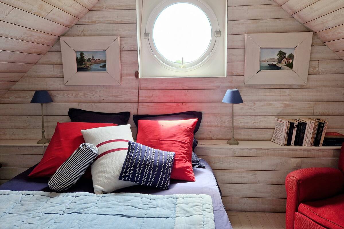 A cozy bedroom with a sloped ceiling, a round skylight, two framed pictures, a bed with red and white pillows, two bedside lamps, and a red chair.