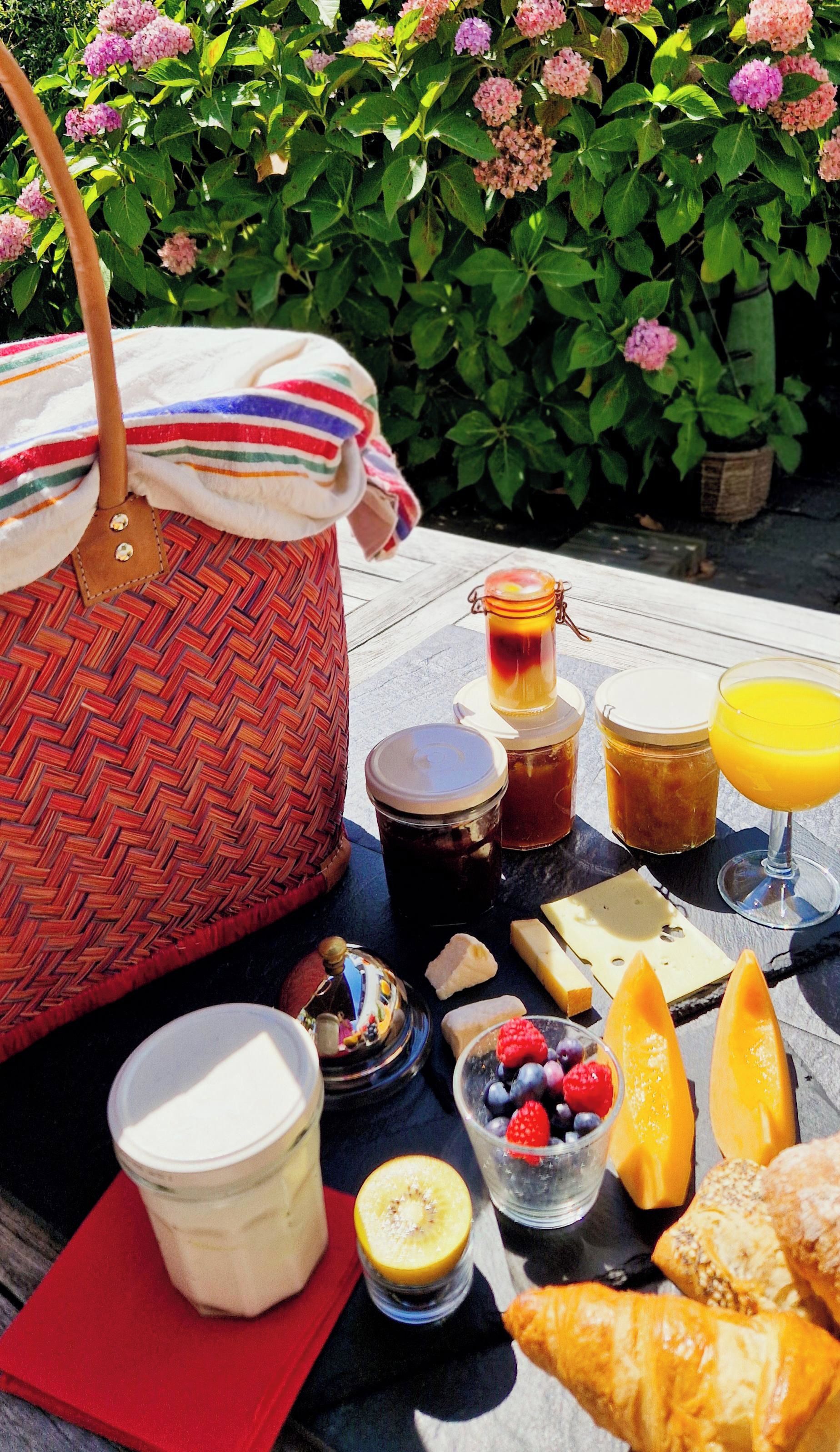 A picnic setup with a woven basket, jars of honey and jam, a glass of orange juice, cheese, fruits, and pastries on a table outdoors.