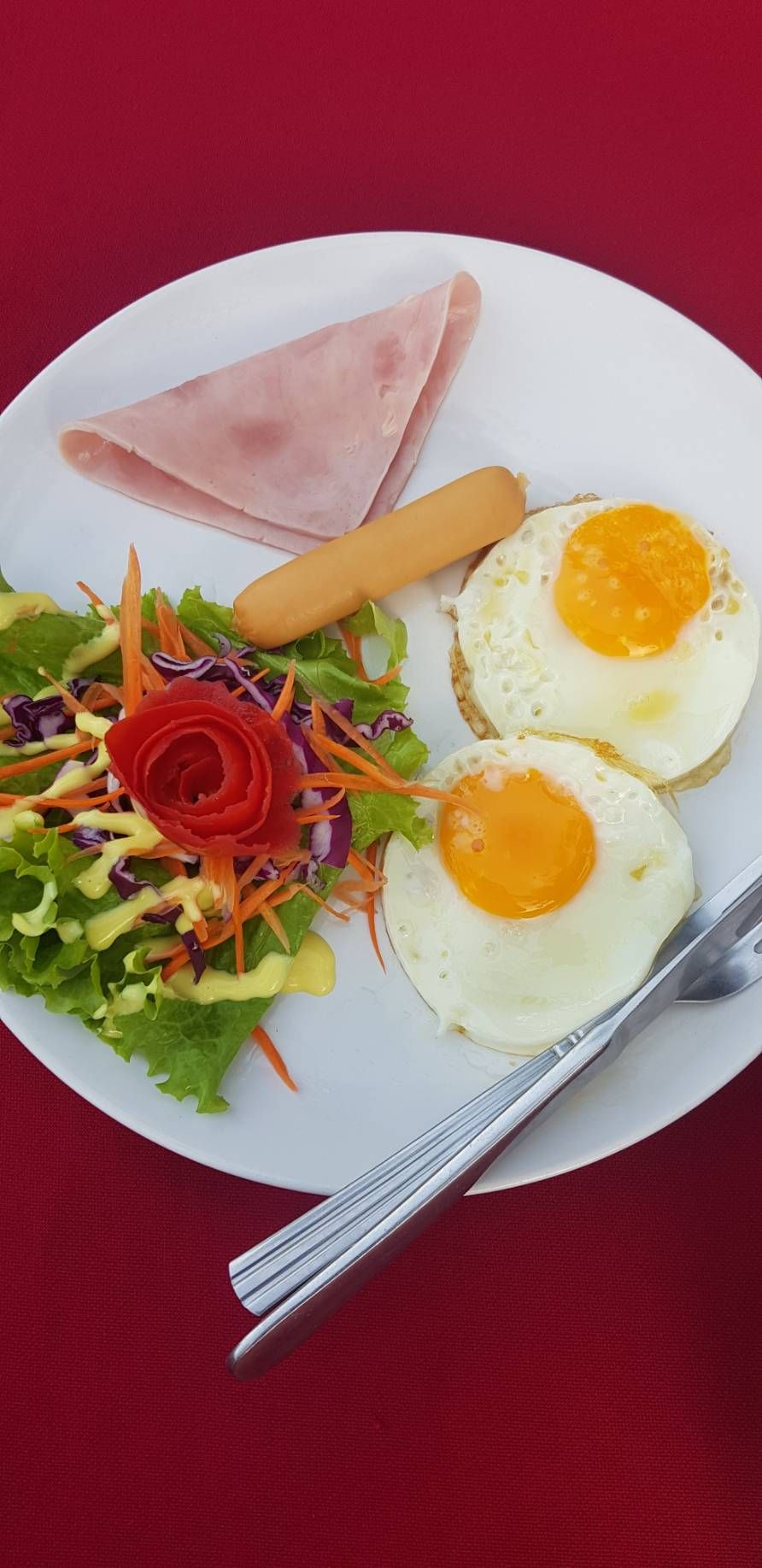 Un plato con dos huevos fritos, una rebanada de jamón y una ensalada con una rosa roja decorativa.