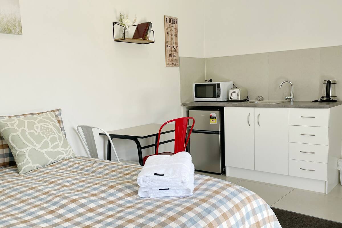 A neatly arranged bedroom with a bed, a chair, a desk, and a kitchenette.