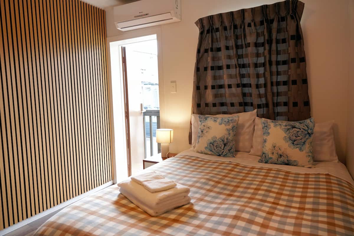 A hotel room with a checkered bedspread, a neatly folded white towel, floral pillows, a window with curtains, and a wooden slat wall.