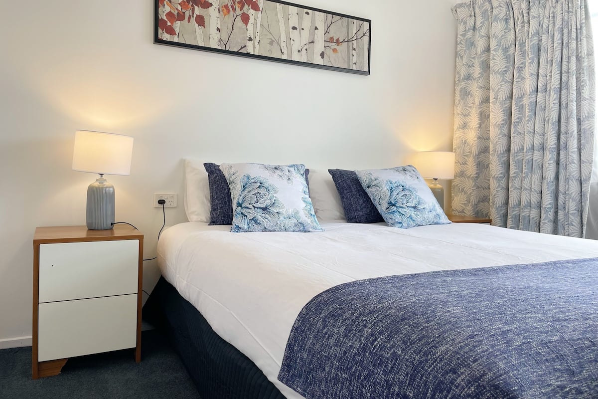 A neatly made bed with a white and blue floral duvet, two matching pillows, a small bedside table with a lamp, and a framed artwork above the bed.