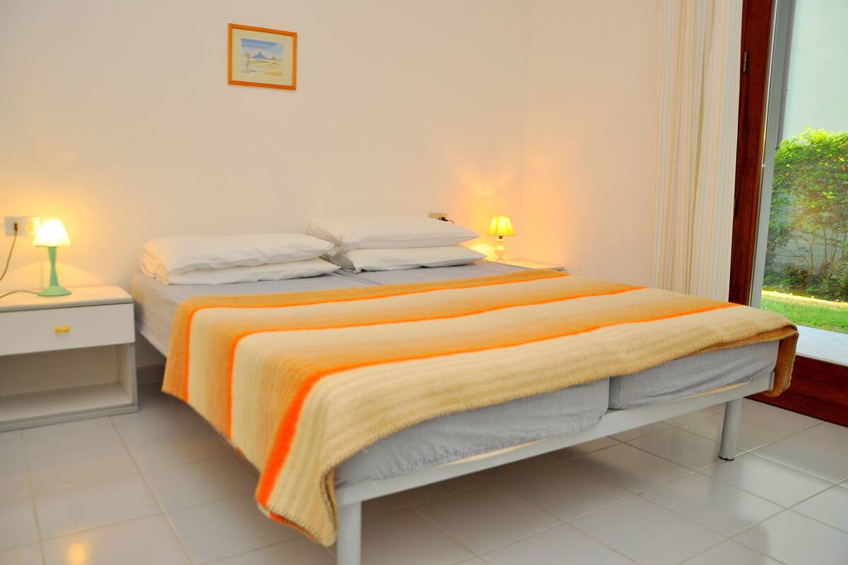 A neatly made bed with white bedding and a striped blanket, a bedside table with a lamp, and a window with curtains.
