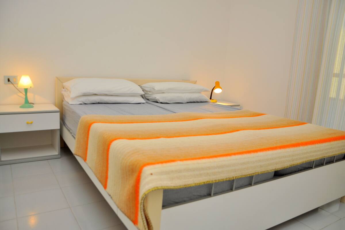 A neatly made bed with a white headboard, two bedside tables with lamps, and a window with sheer curtains.