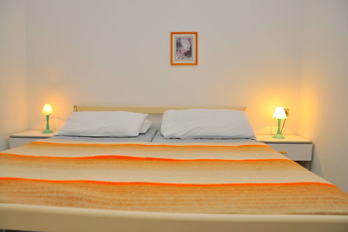 A neatly made bed with white linens and an orange blanket, flanked by two bedside tables with lamps.