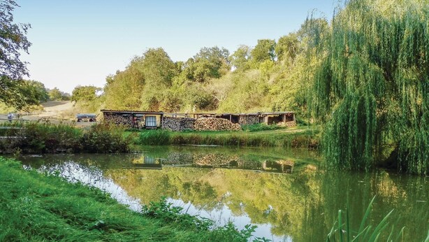 Un chalet rustique situé au bord d’un étang, dont l’image se reflète dans l’eau, et entouré d’arbres et de verdure.