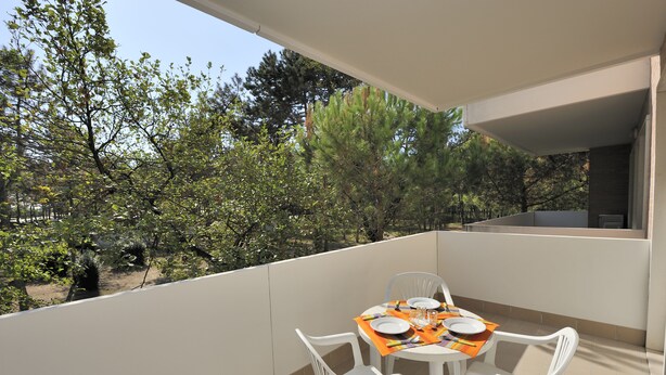 A balcony with a table set for two, surrounded by white chairs, overlooking a garden with trees and shrubs. - Residence Gemini (San Michele al Tagliamento)