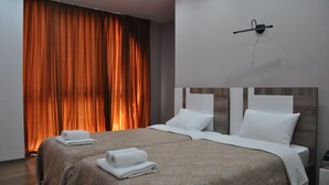 Un lit double avec deux oreillers blancs et une couvre-lit beige, une table de chevet avec une lampe, et une grande fenêtre avec des rideaux orange.