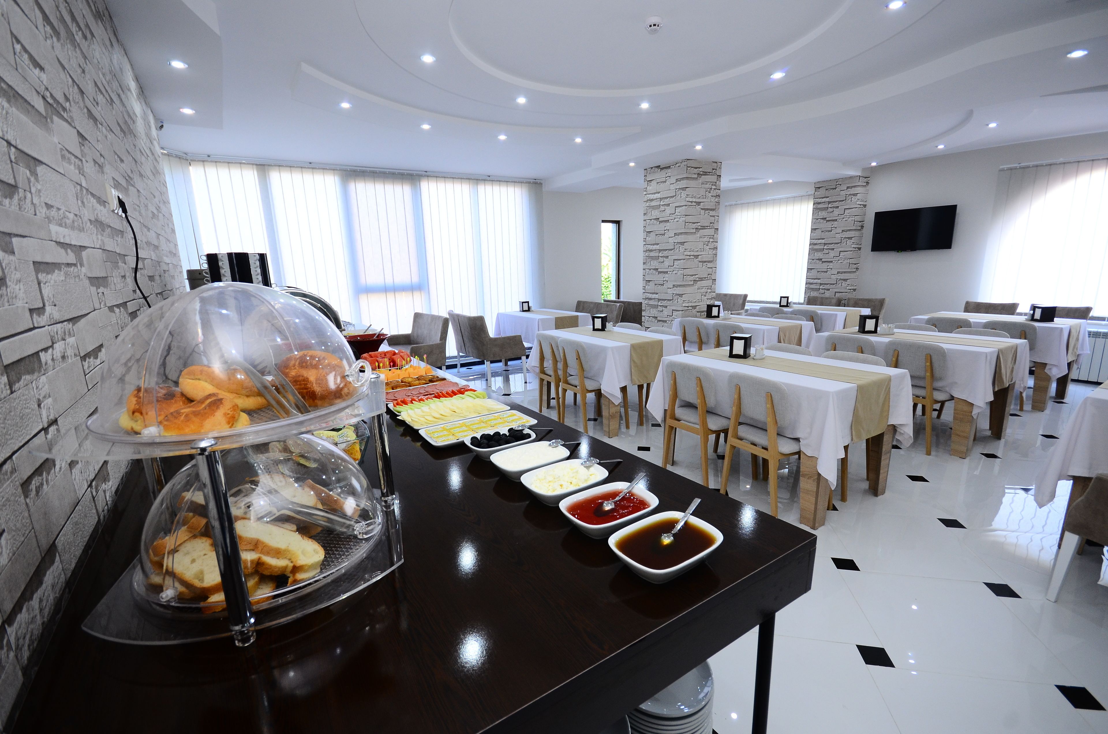 A buffet setup with various food items and a clear glass dome covering some pastries. Tables are arranged in the background with a stone wall and a flat-screen TV mounted on the wall.