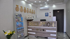 Un comptoir d’accueil avec un revêtement en bois et une enseigne indiquant « RÉCEPTION ». On peut y voir des horloges au mur, un panneau d’affichage avec des brochures et un vase rempli de fleurs sur le comptoir.