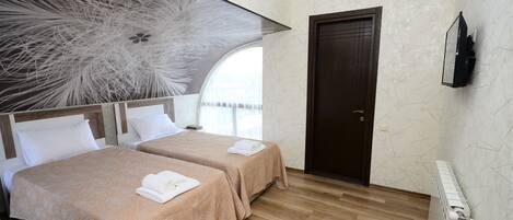 A hotel room with two beds, a large abstract artwork on the wall, a wooden floor, a dark door, and a mounted TV.