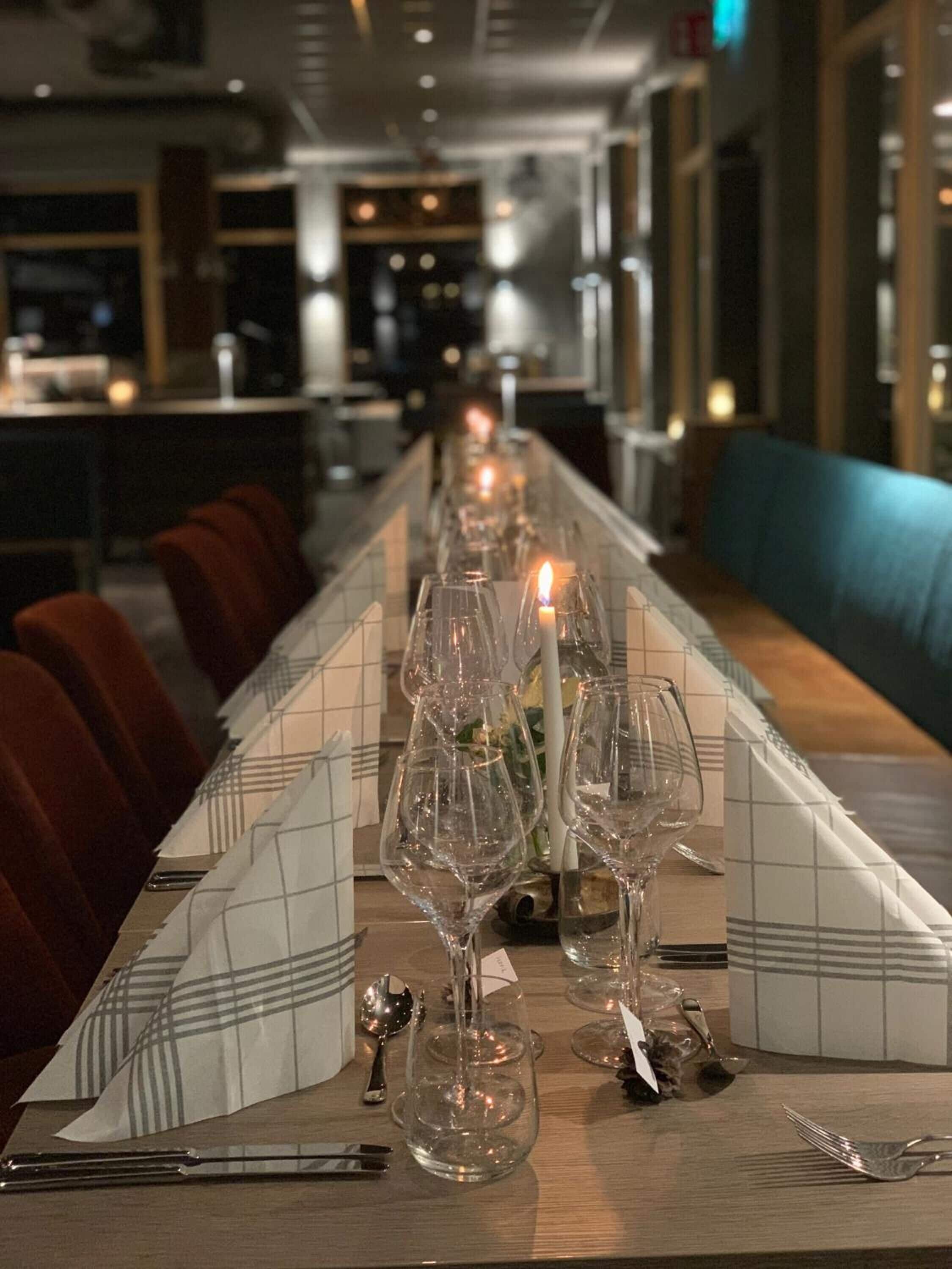 Une longue table à manger, ornée de nombreux verres à vin et d’une bougie allumée, est placée dans un restaurant à la lumière tamisée, dont l’intérieur est de style moderne.