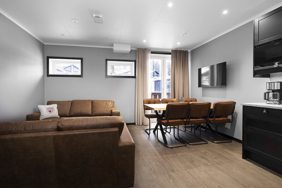 A modern living room with a brown leather sofa, a dining table with four chairs, and a kitchen area with a black refrigerator and a coffee machine.