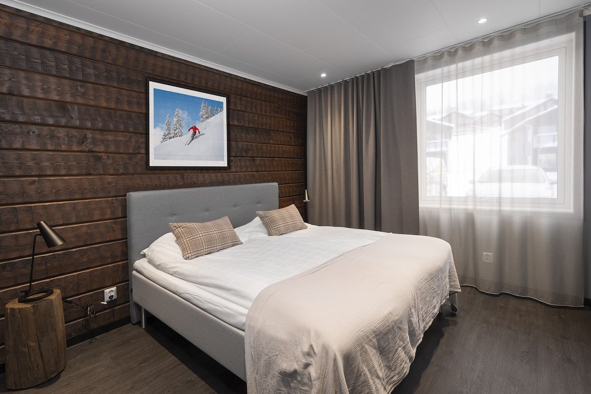 A modern bedroom with a wooden wall, a large bed, a framed picture, and a window with curtains.