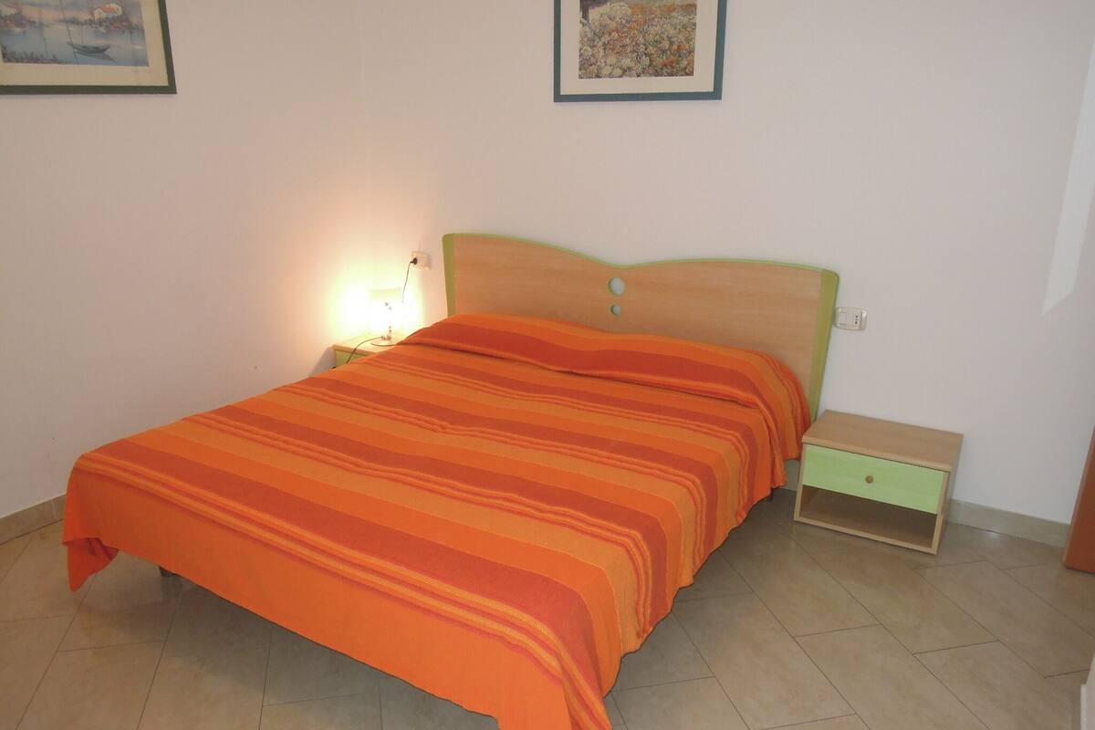 Un letto con copriletto a righe arancioni, una testiera in legno, un comodino con una lampada e un piccolo mobiletto con cassetti.