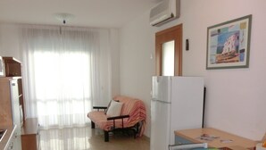 Un espacio habitable compacto con kitchenette, un comedor con sillas azules, un sofá con una manta a rayas y un ventanal con cortinas translúcidas.