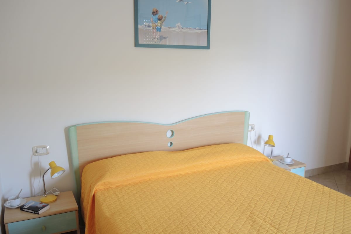 Un letto con una testiera in legno, una coperta trapuntata gialla, un comodino con una lampada e un quadro appeso al muro.