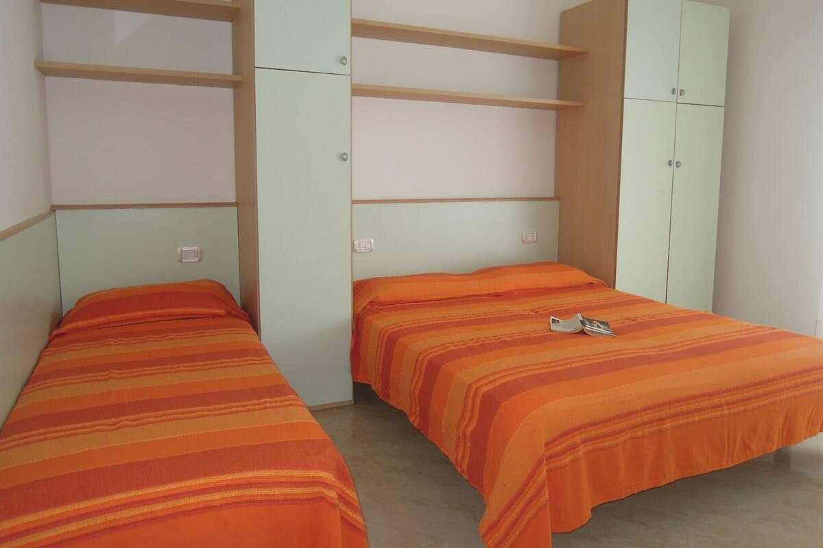 Una stanza con due letti, ognuno con lenzuola a righe arancioni, e armadi verde chiaro con ripiani multipli.