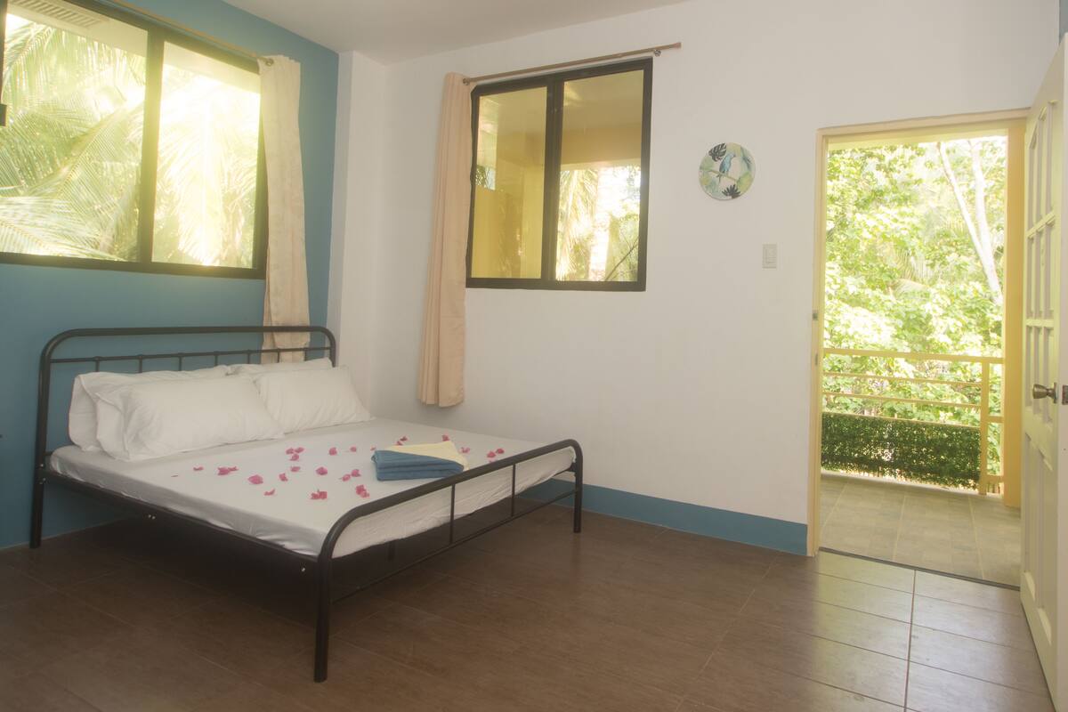 A bedroom with a single bed, a window with curtains, a mirror, and a door leading to a balcony.