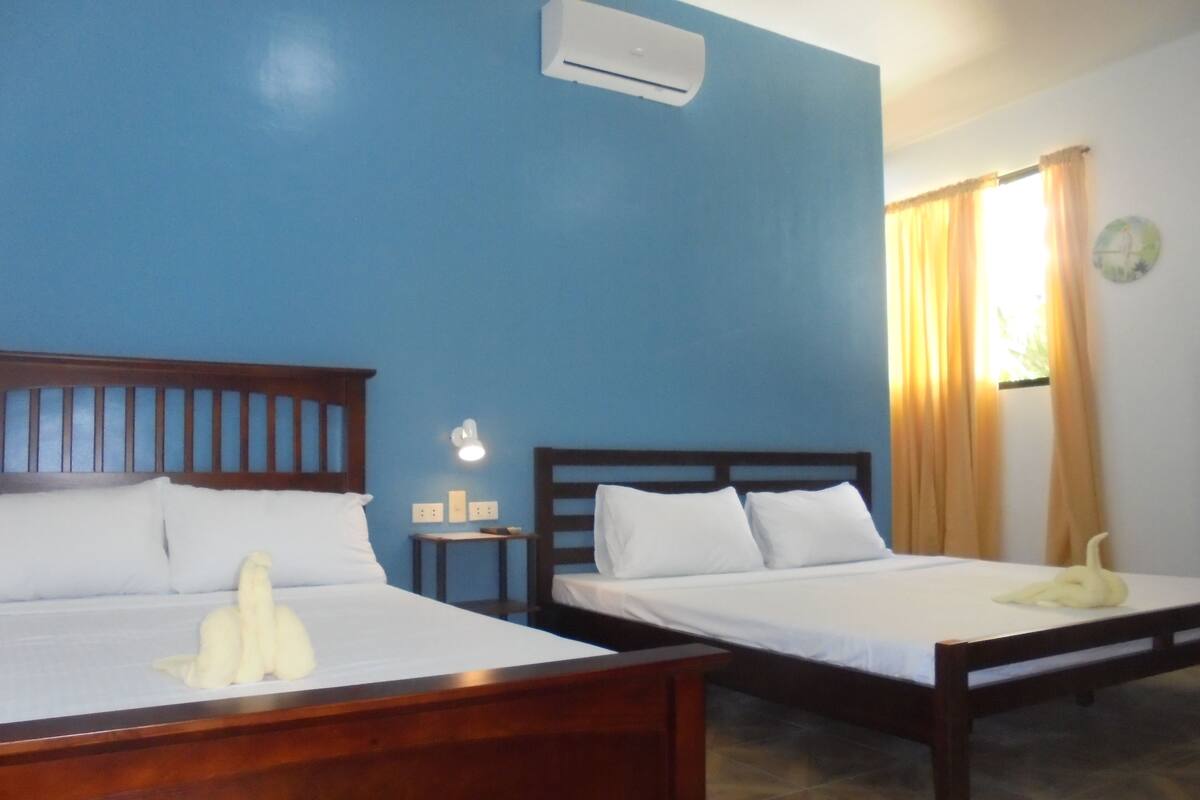 A hotel room with two wooden beds, a wall-mounted air conditioner, a small bedside table with a lamp, and a window with curtains.