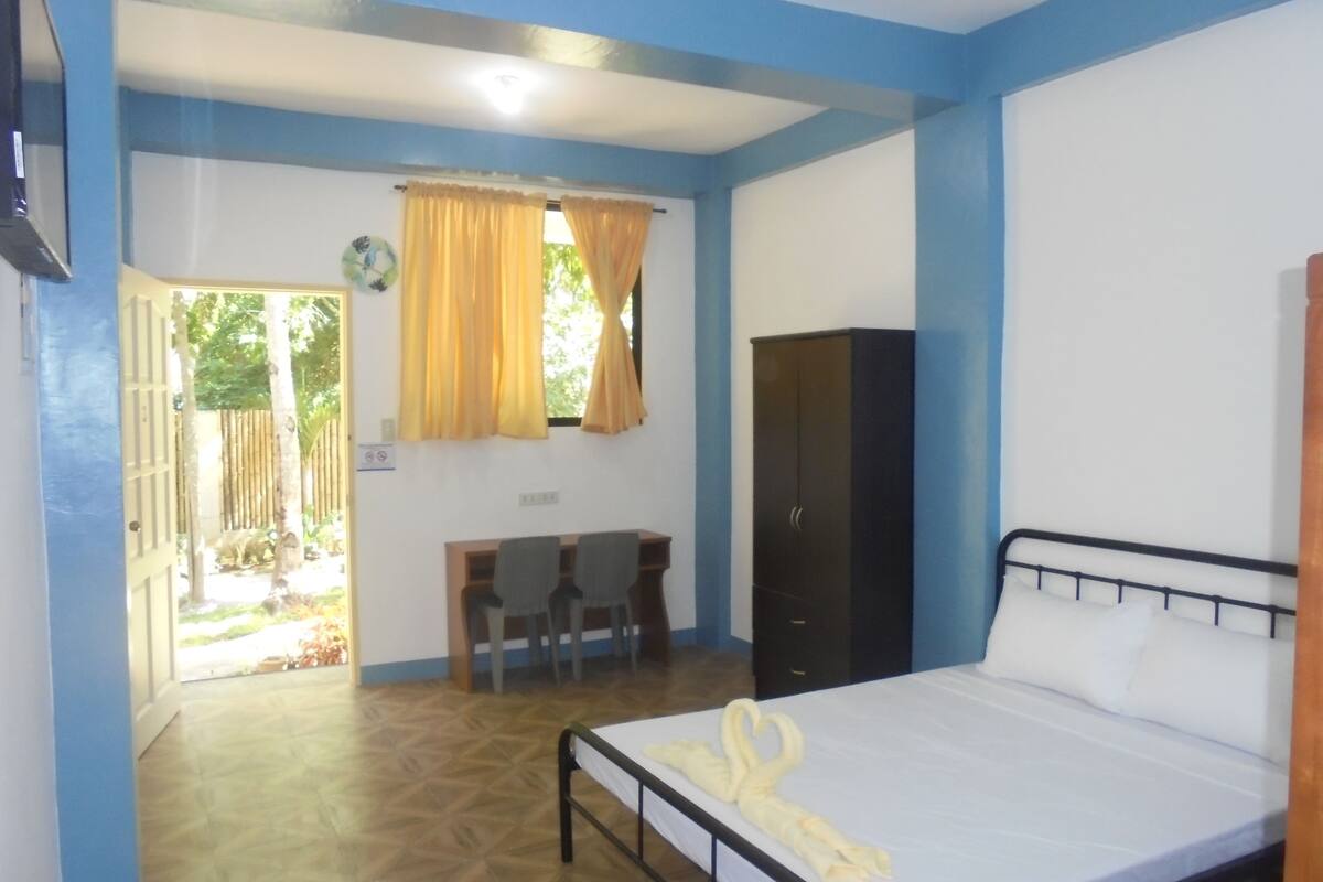 A bedroom with a bed, a wardrobe, a desk with chairs, and a window with curtains.