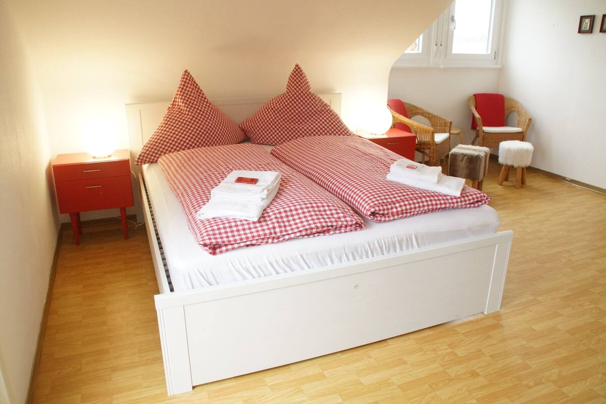A neatly made bed with checkered bedding, a red bedside table, a chair, and a small stool.