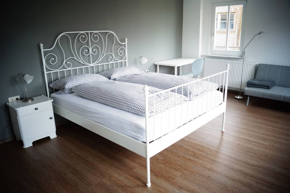 A neatly arranged bedroom with a metal bed frame, a bedside table with a lamp, a small desk, and a grey sofa.