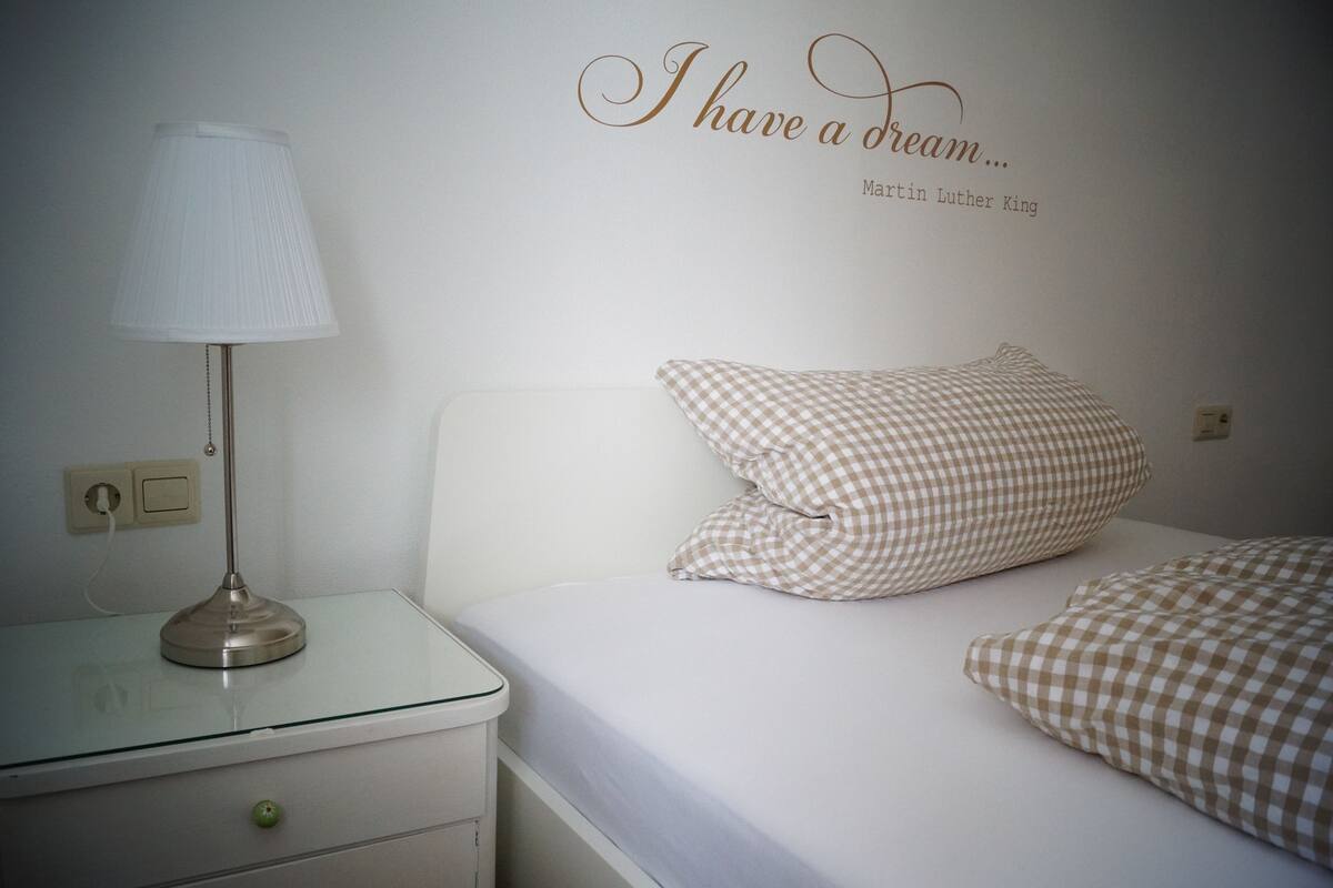 A bedroom with a checkered pillow, a bedside lamp, and a wall quote.