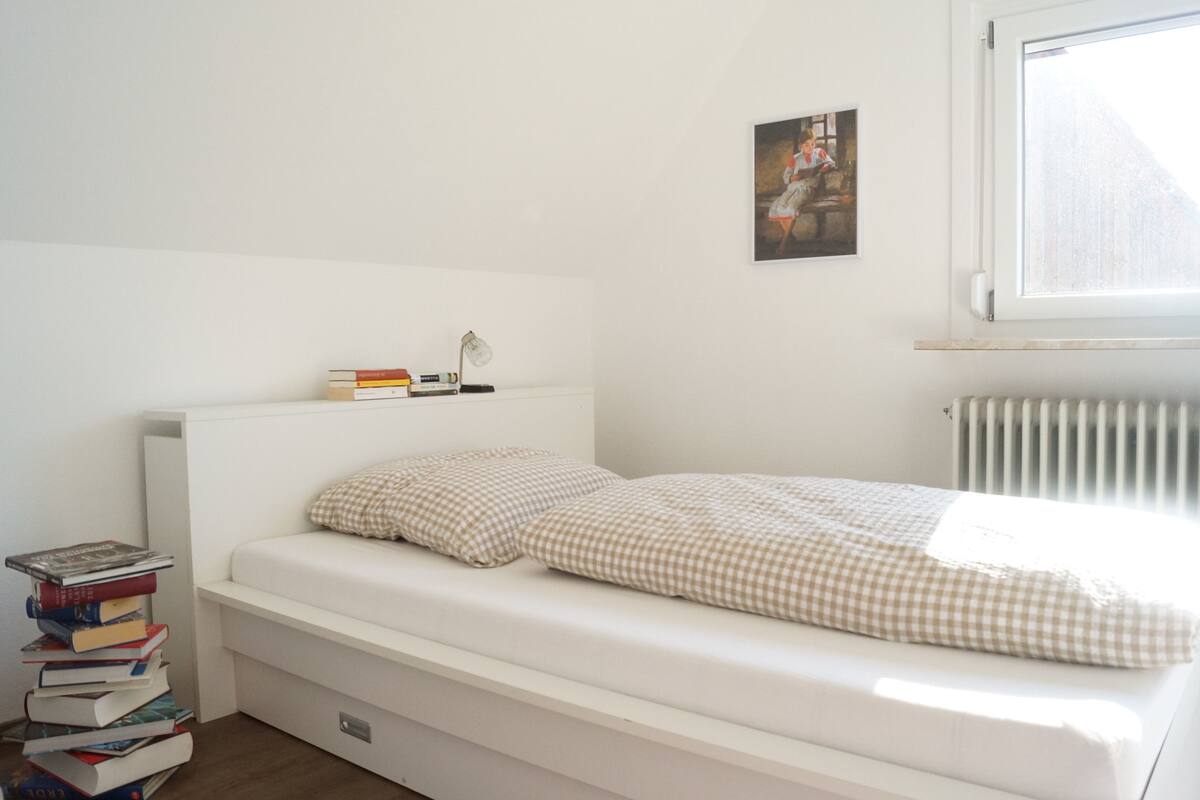 A small, neatly made bed with a checkered bedspread and pillows, a stack of books beside it, a lamp on a bedside table, and a framed picture on the wall.