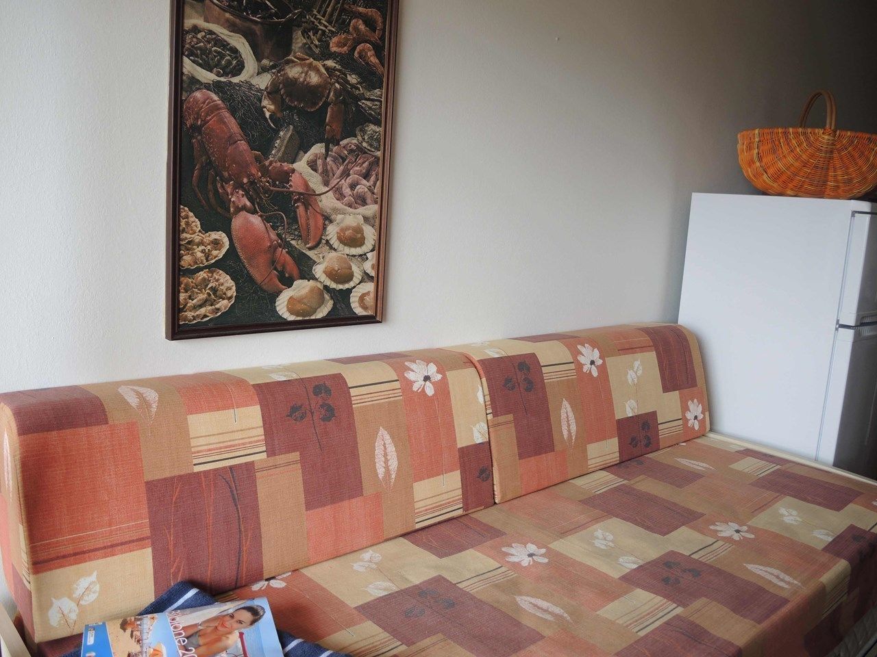 Ein Sofa mit floralem und geometrischem Muster, ein gerahmtes Bild von Meeresfrüchten an der Wand und ein Korbwäschekorb auf einem Schrank.