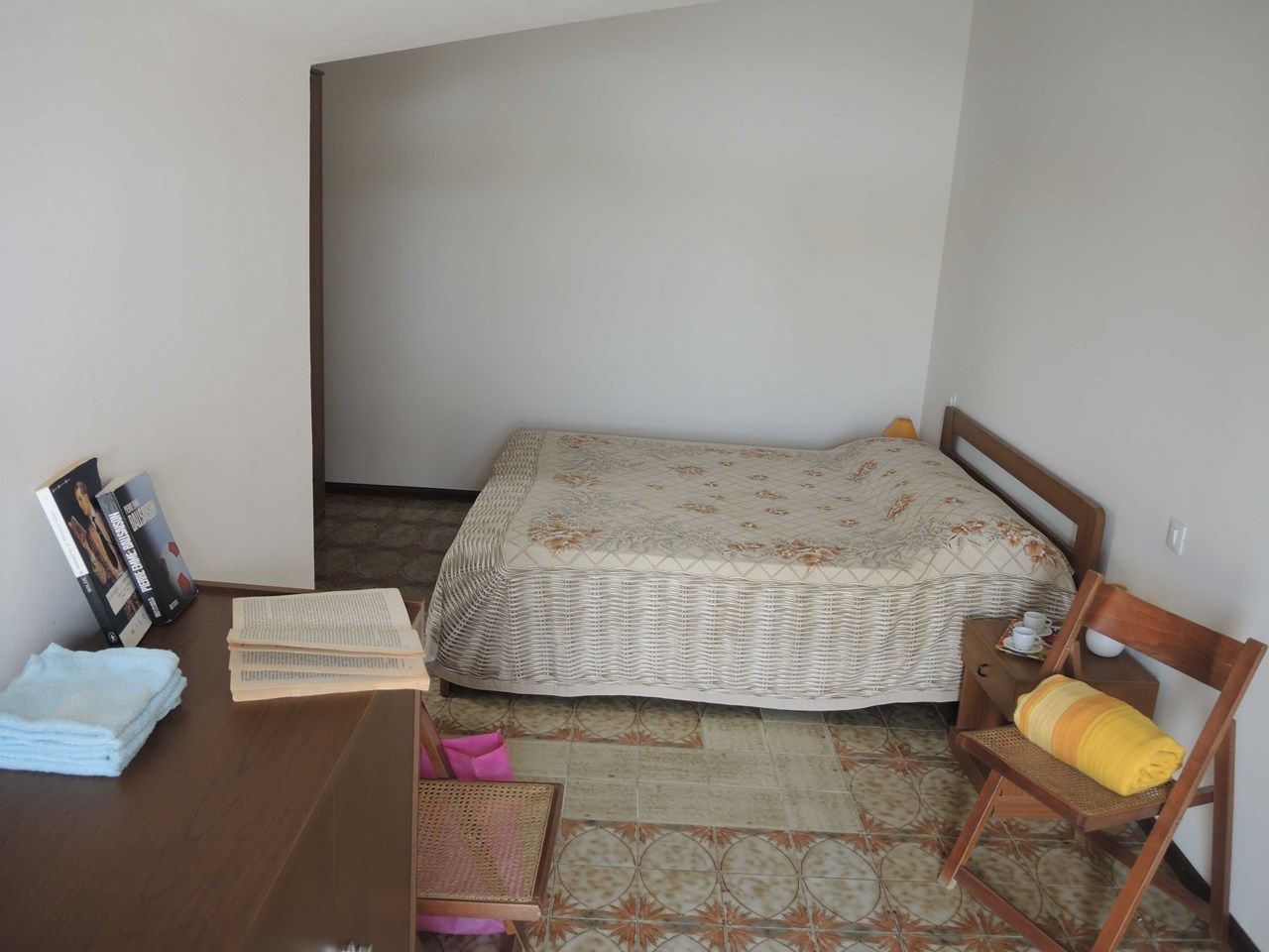 A small, neatly arranged bedroom with a single bed, a wooden desk with books, a chair, and a small table with a cup and saucer.