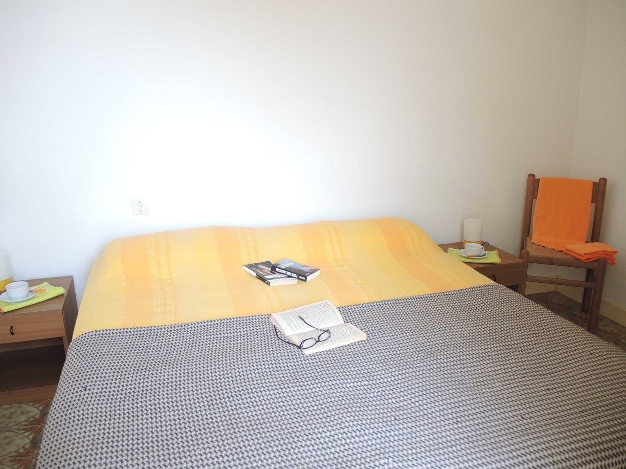 A neatly made bed with a patterned bedspread, a pair of glasses, and a book on it. There is a chair with an orange towel beside it, and a small table with a cup and a book.