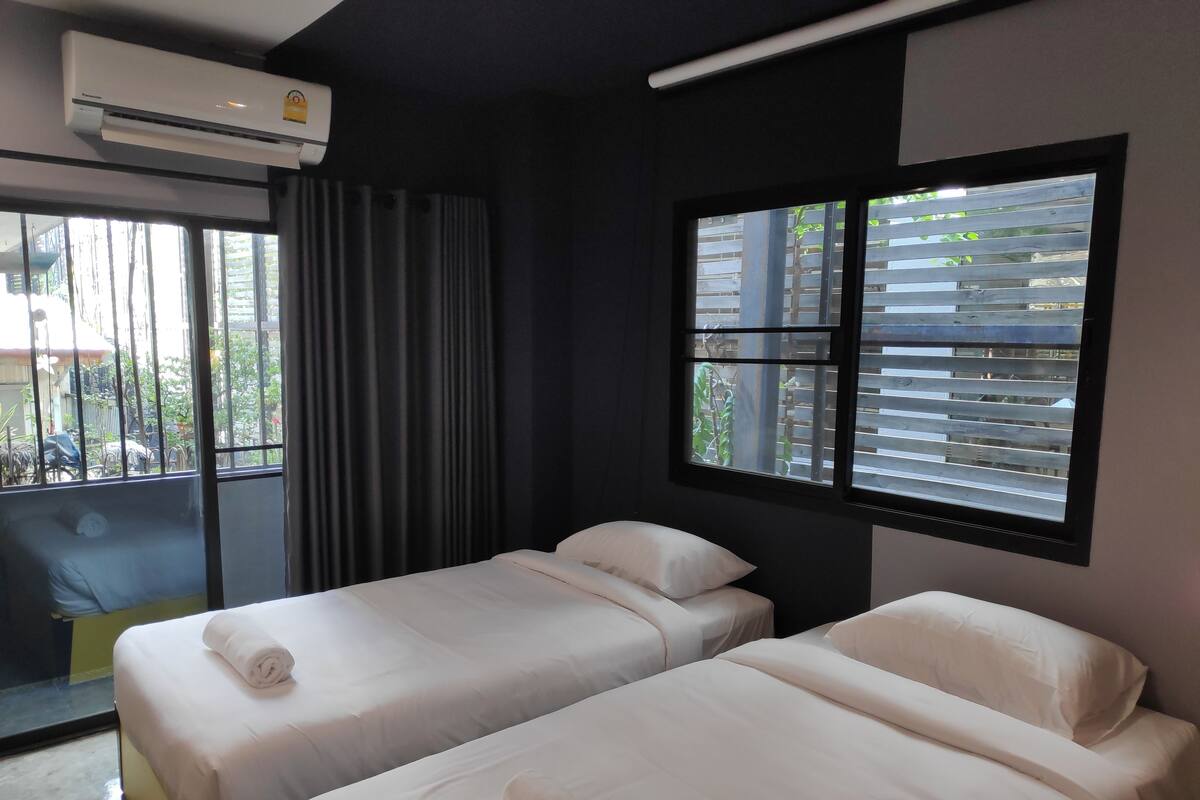 A hotel room with two beds, a large window with blinds, an air conditioning unit, and a view of the outside.