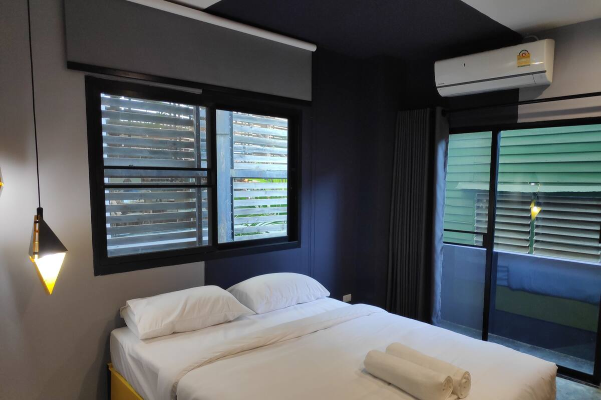 A hotel room with a bed, a window with blinds, an air conditioning unit, and a hanging light fixture.