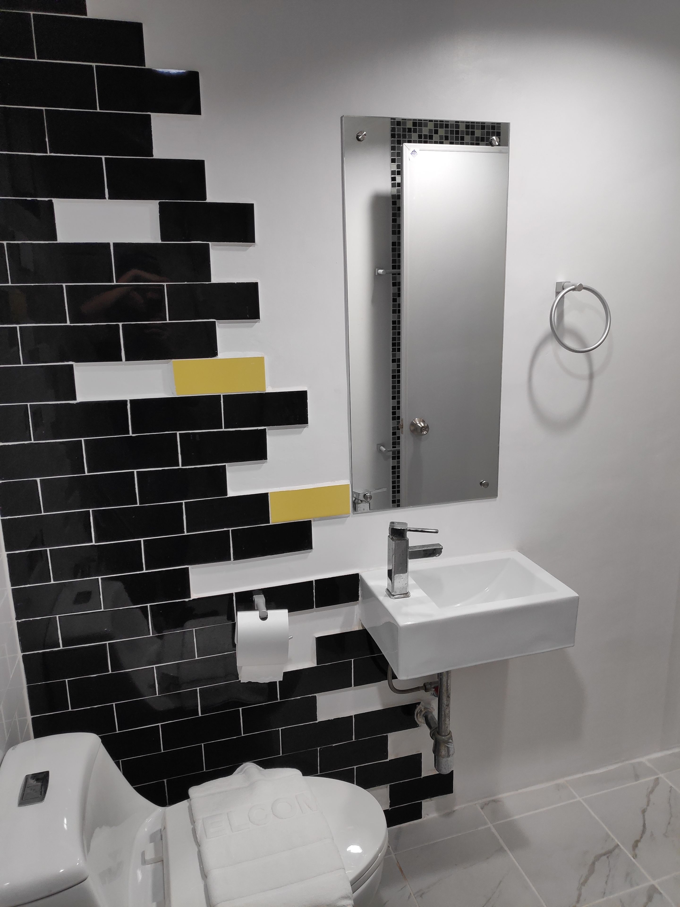 Un baño con un lavamanos blanco, un espejo y un inodoro. La pared tiene azulejos blanco y negro con detalles amarillos.