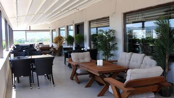 Een overdekt terras met tafels, stoelen, plantenbakken en grote ramen met uitzicht.