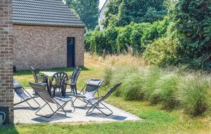 Een bakstenen gebouw met een pannendak, een terras met een tafel en stoelen, en een gazon met hoge planten.