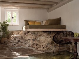 Quarto aconchegante com uma cama embutida em uma parede de pedra texturizada, uma mesa de madeira com um vaso de uvas verdes e uma cadeira de vime.