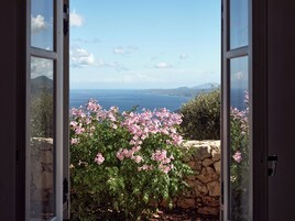 Vista a través de una puerta abierta que muestra un muro de piedra con flores rosadas y una escalera de piedra que conduce a un balcón con vista a un cuerpo de agua y montañas distantes.