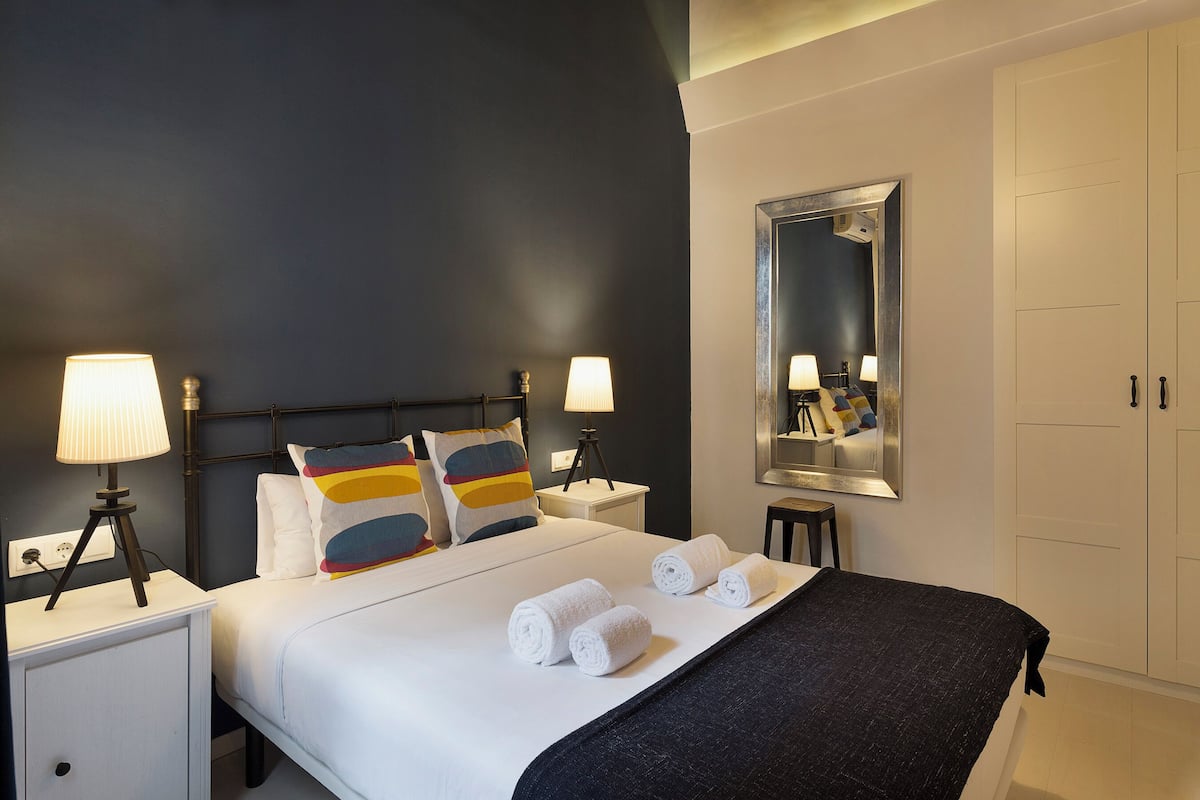 Un lit bien fait, avec du linge de lit blanc et des serviettes pliées, une lampe de chevet, une table de chevet, un miroir et une armoire.