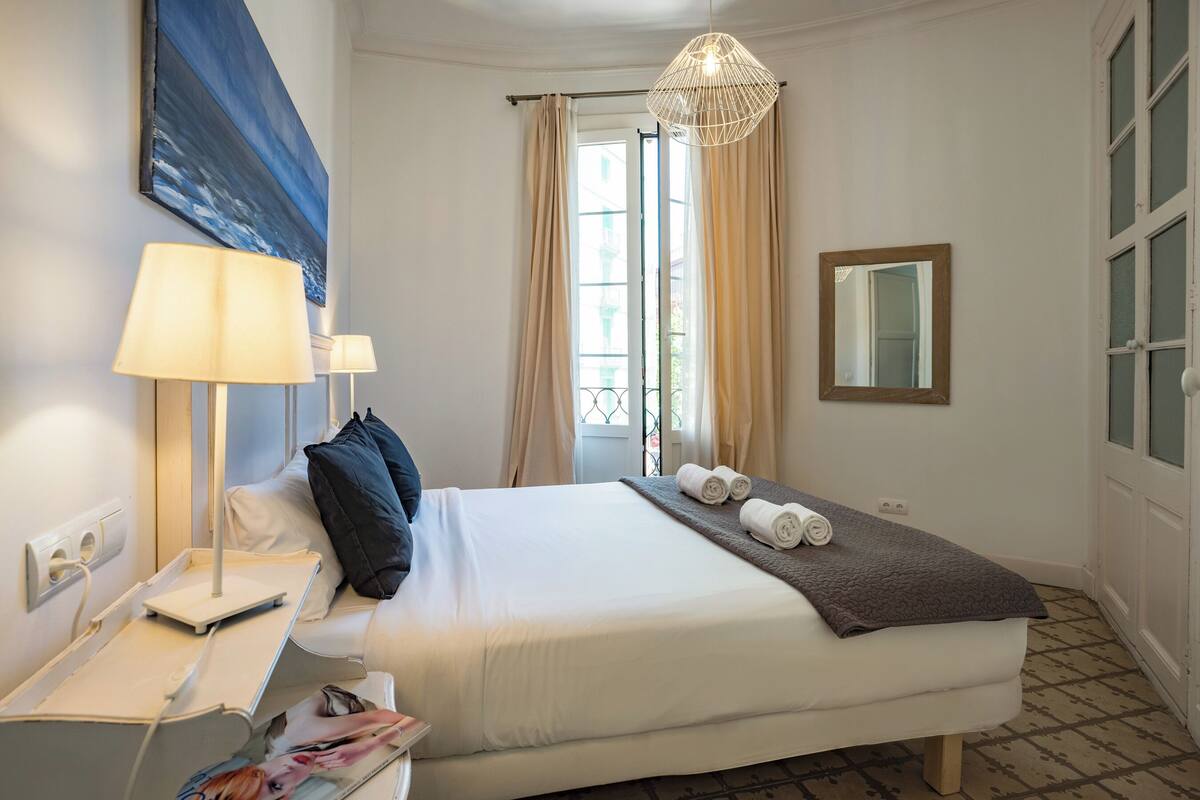 Une chambre d’hôtel comprenant un lit, deux lampes de chevet, une fenêtre avec des rideaux, un luminaire suspendu et un miroir fixé au mur.