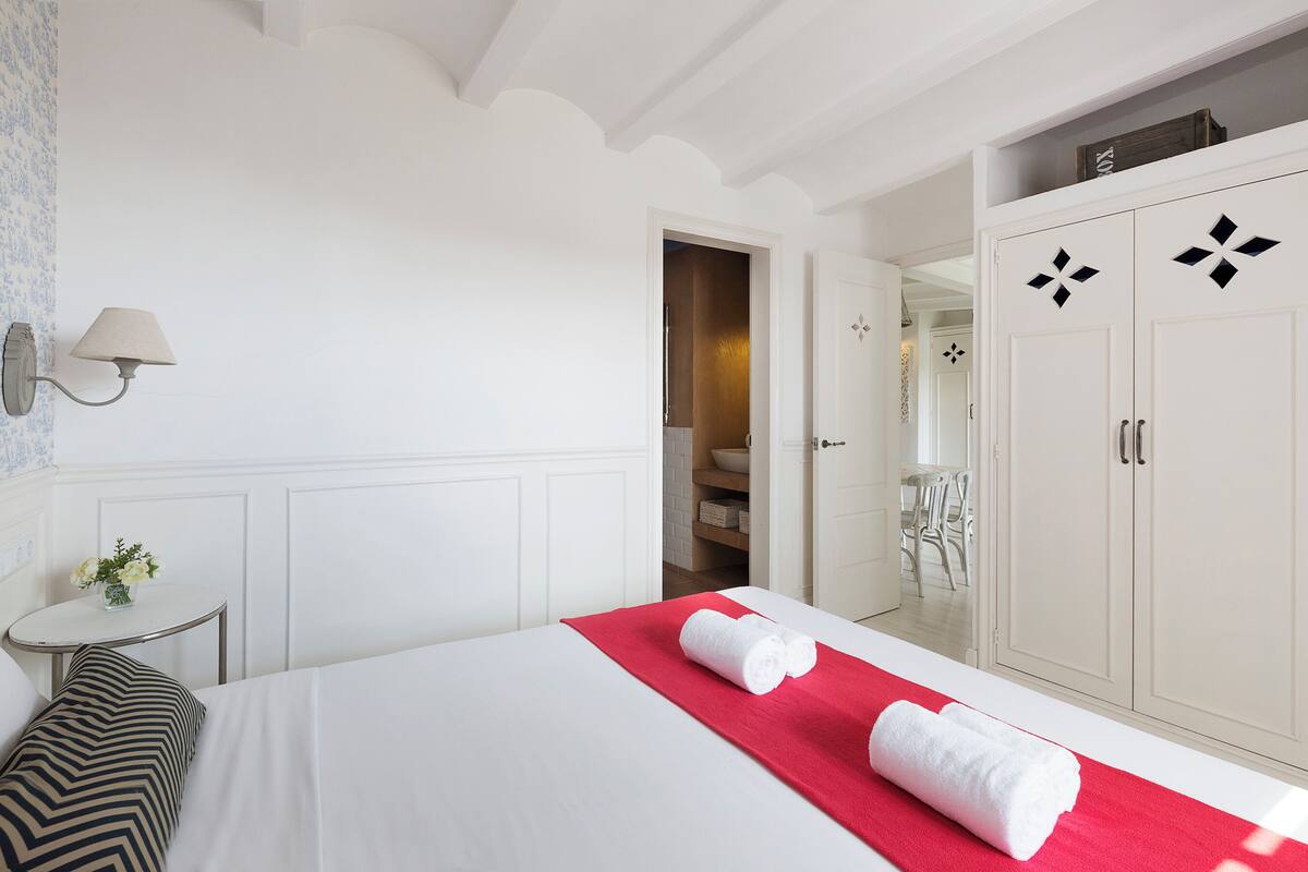 Une chambre d’hôtel avec un lit soigneusement fait, une petite table ronde avec une lampe et une armoire ornée de motifs décoratifs.