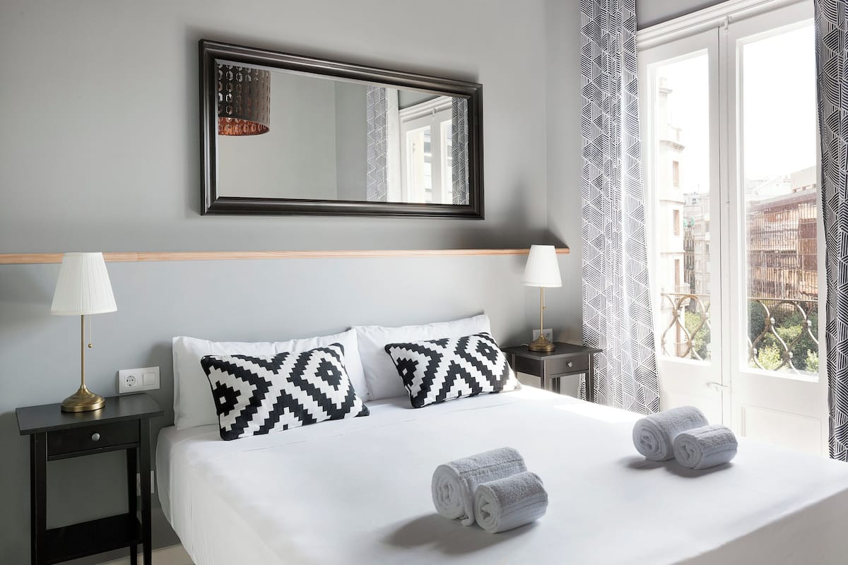 Une chambre d’hôtel avec un lit, deux lampes de chevet, un miroir et une fenêtre avec des rideaux.