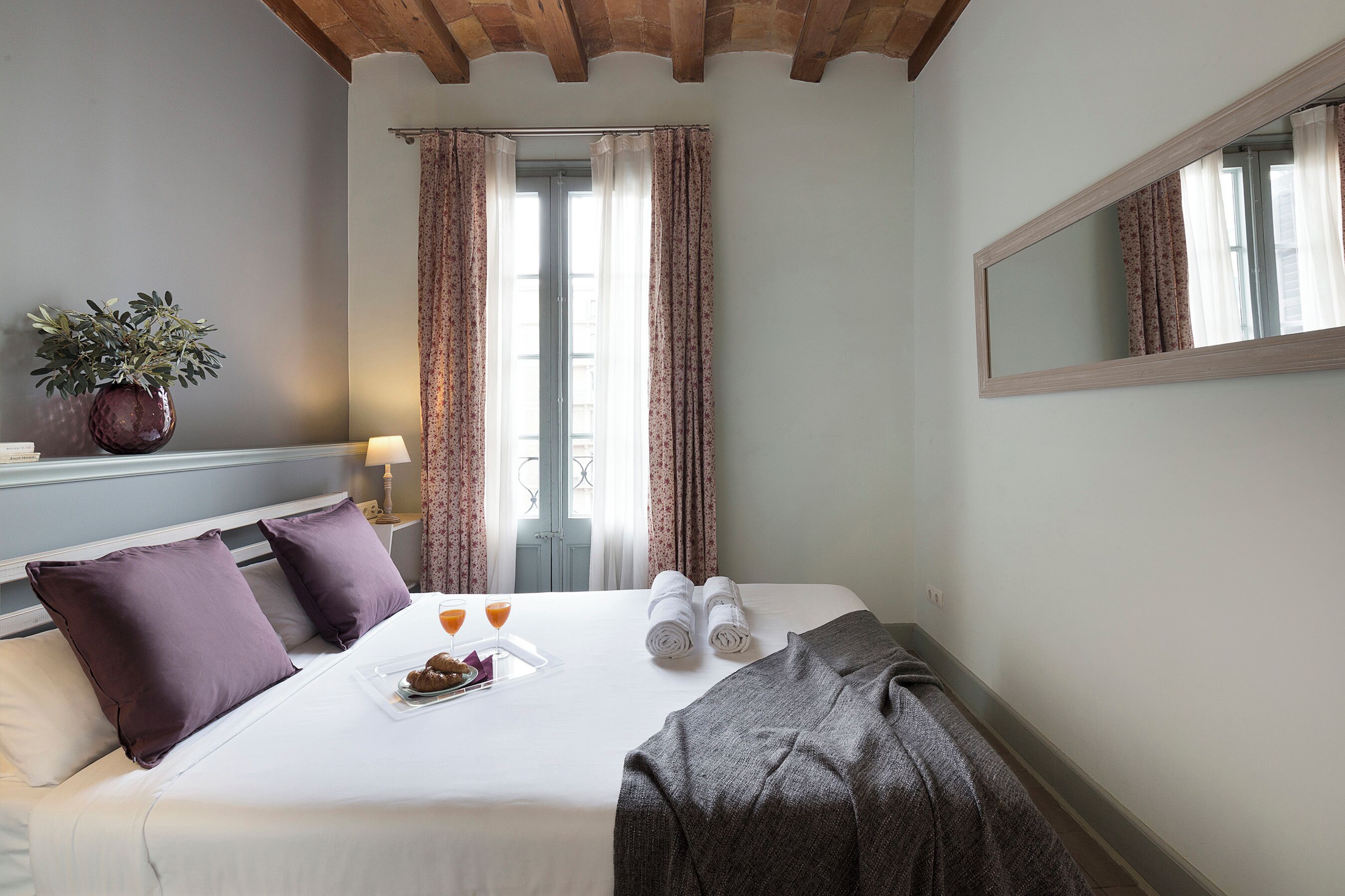 Une chambre d’hôtel avec un lit, des oreillers, un plateau avec de la nourriture et des boissons, une plante et une fenêtre avec des rideaux.