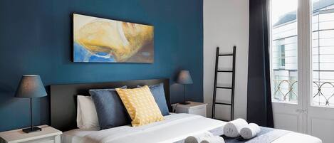 A modern hotel room with a bed, a painting, a nightstand, a lamp, and a window with a view of buildings.