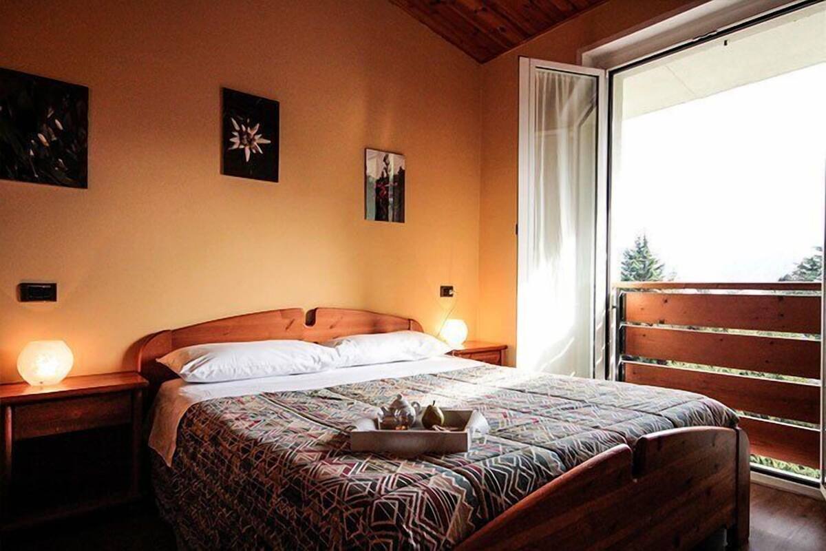 A bedroom with a wooden bed, a bedside table with a lamp, and a window with a view of greenery.