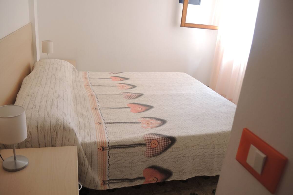 Un lit bien fait, recouvert d’une couvre-lit à motifs, une lampe de chevet et un interrupteur fixé au mur.